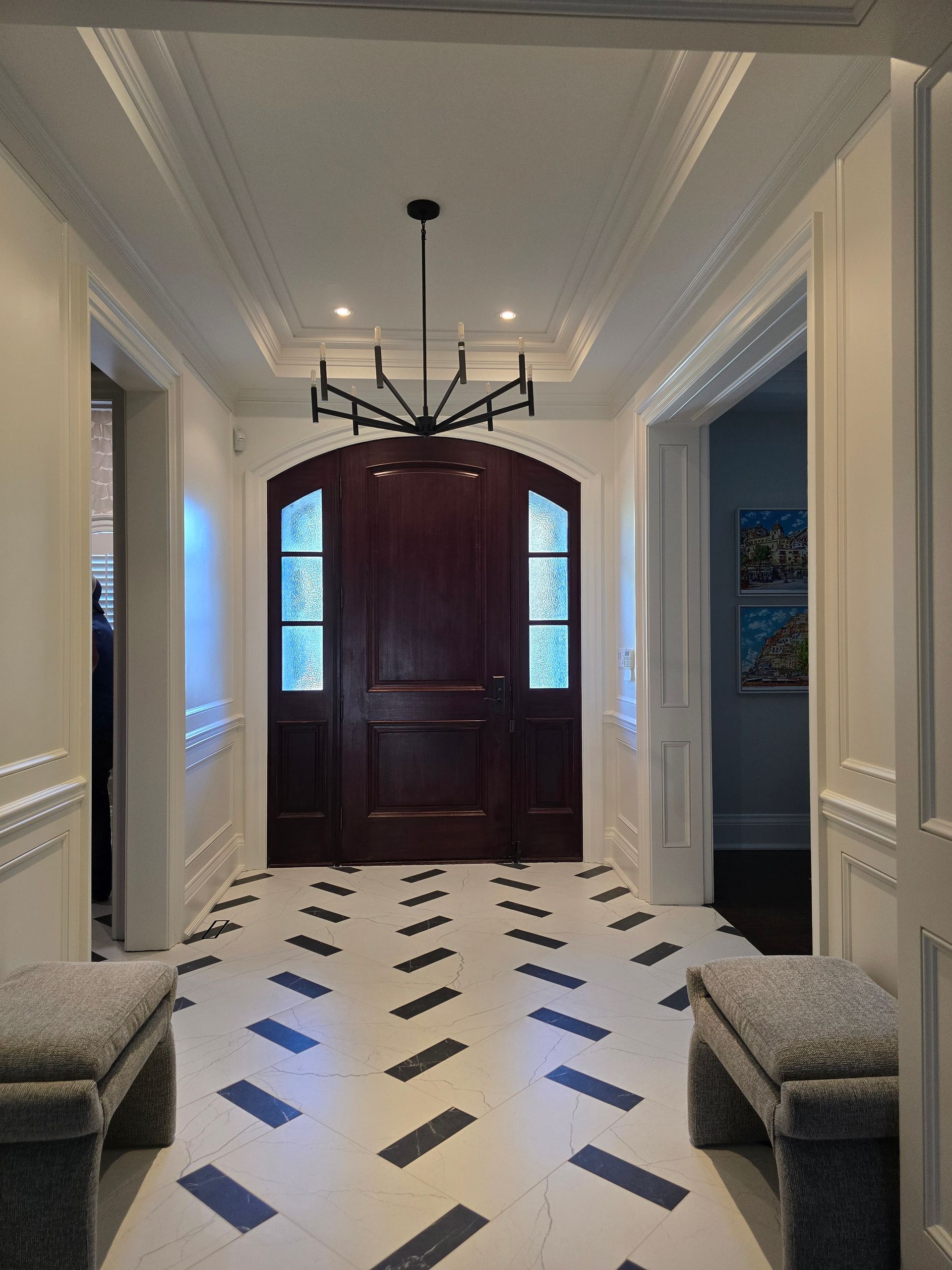 Hallway with dark wooden door, patterned floor, two benches, and chandelier.
