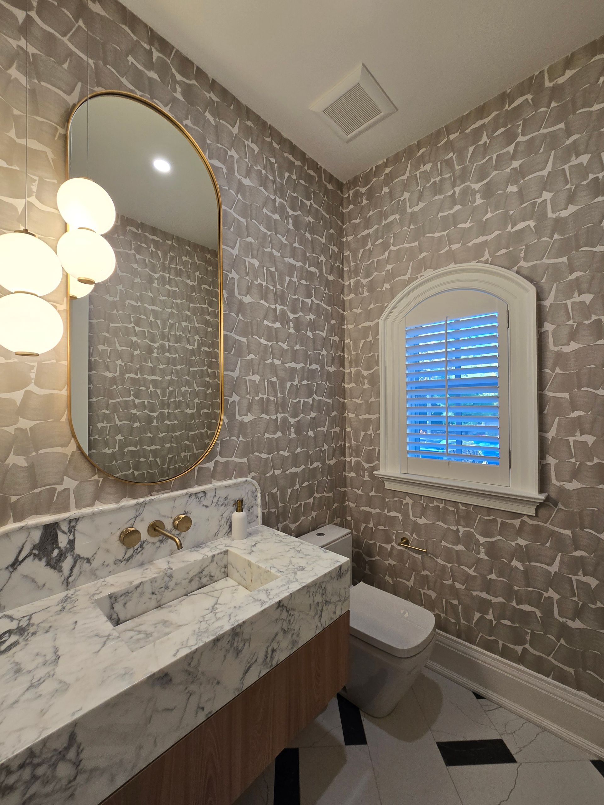 Bathroom with marble sink, gold fixtures, and patterned wallpaper. Oval mirror, globe lights, and arched window.