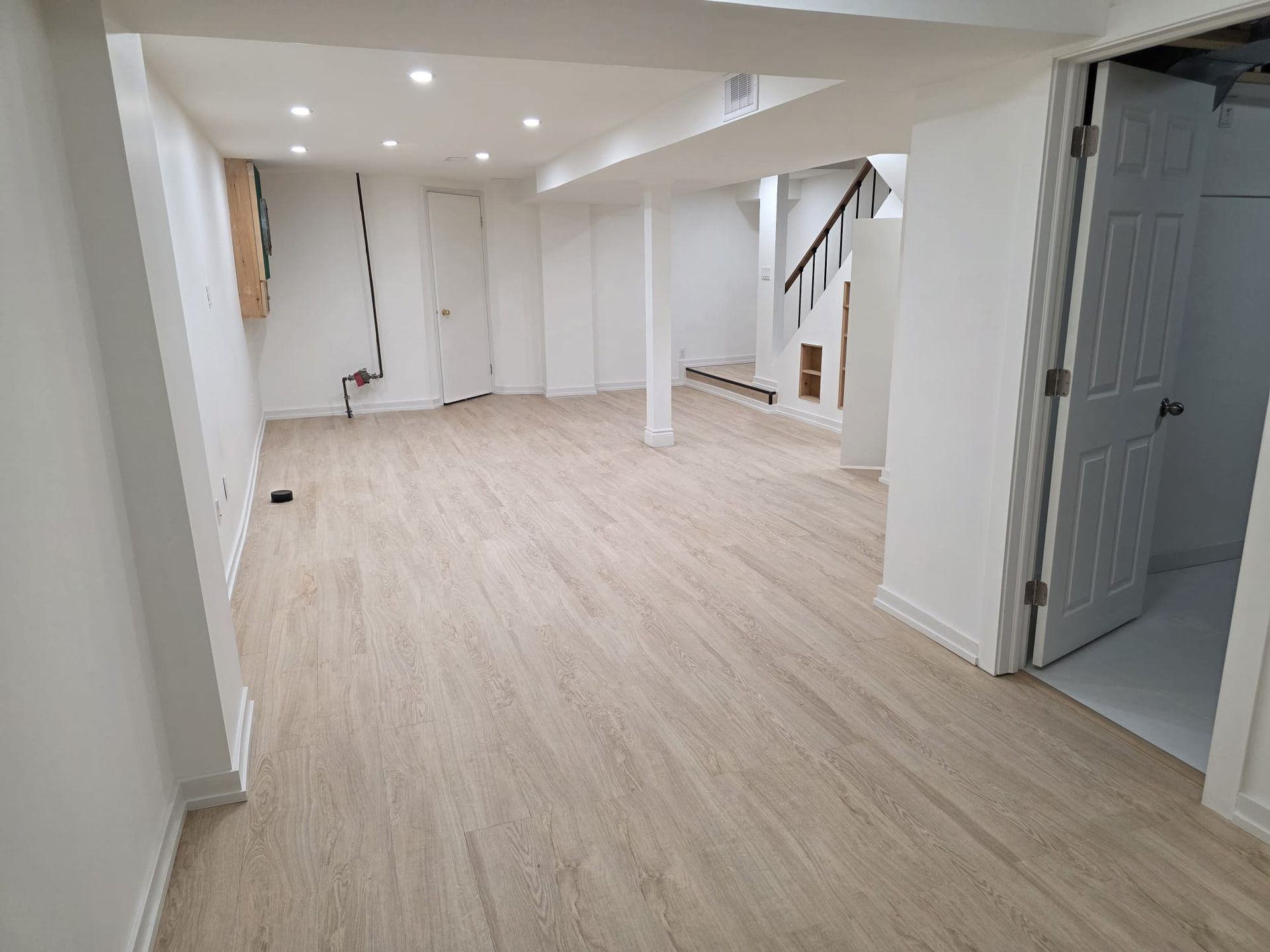 Empty white basement room with light wood flooring, recessed lights, and an open doorway to a gray stairwell