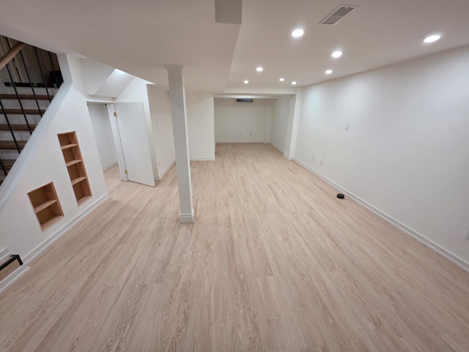 Empty finished basement with light wood floors, white walls, recessed lights, and built-in under-stair shelves.