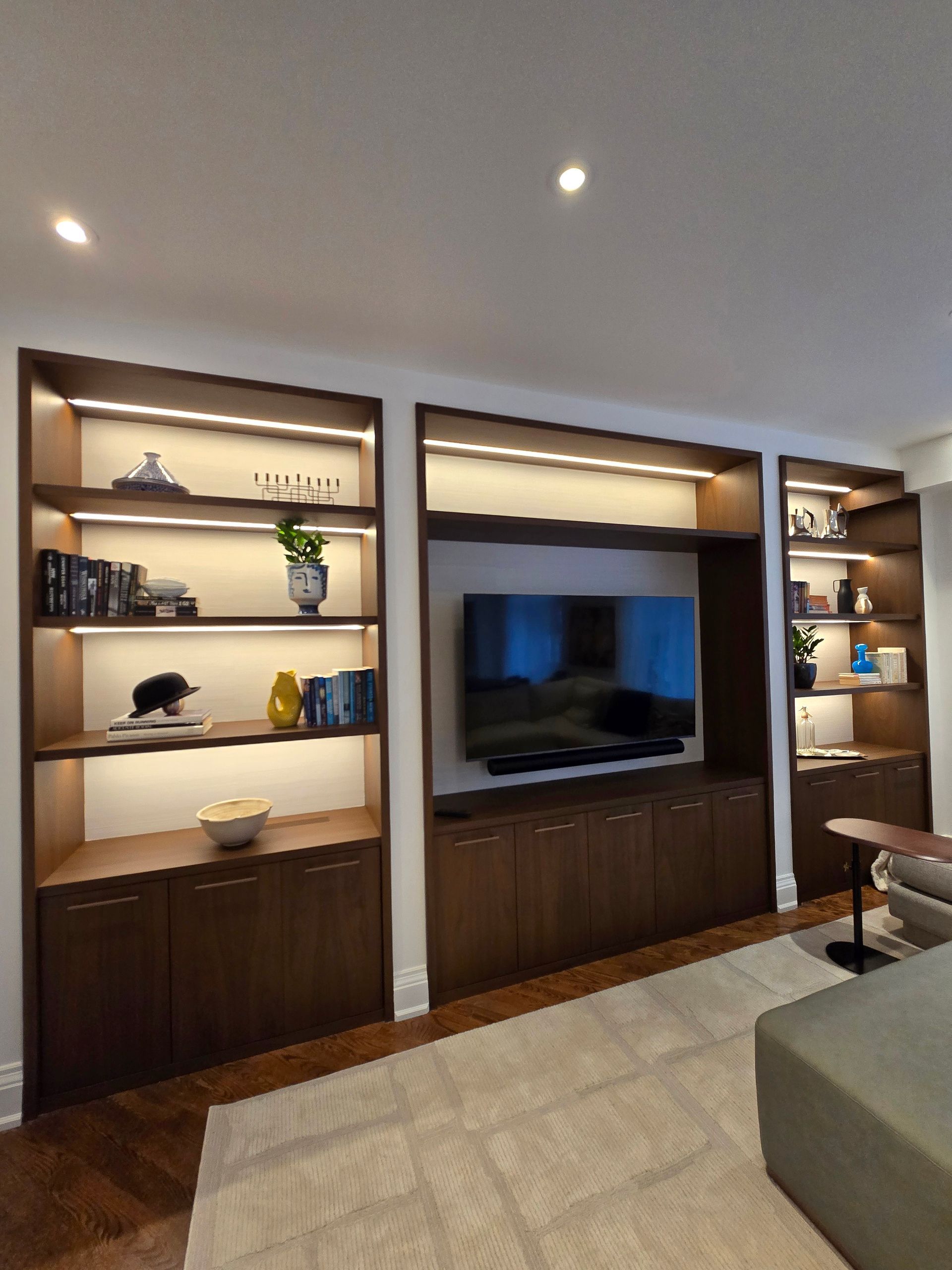Modern living room with built-in wooden shelving and a large TV, warm recessed lighting, and a beige sofa edge