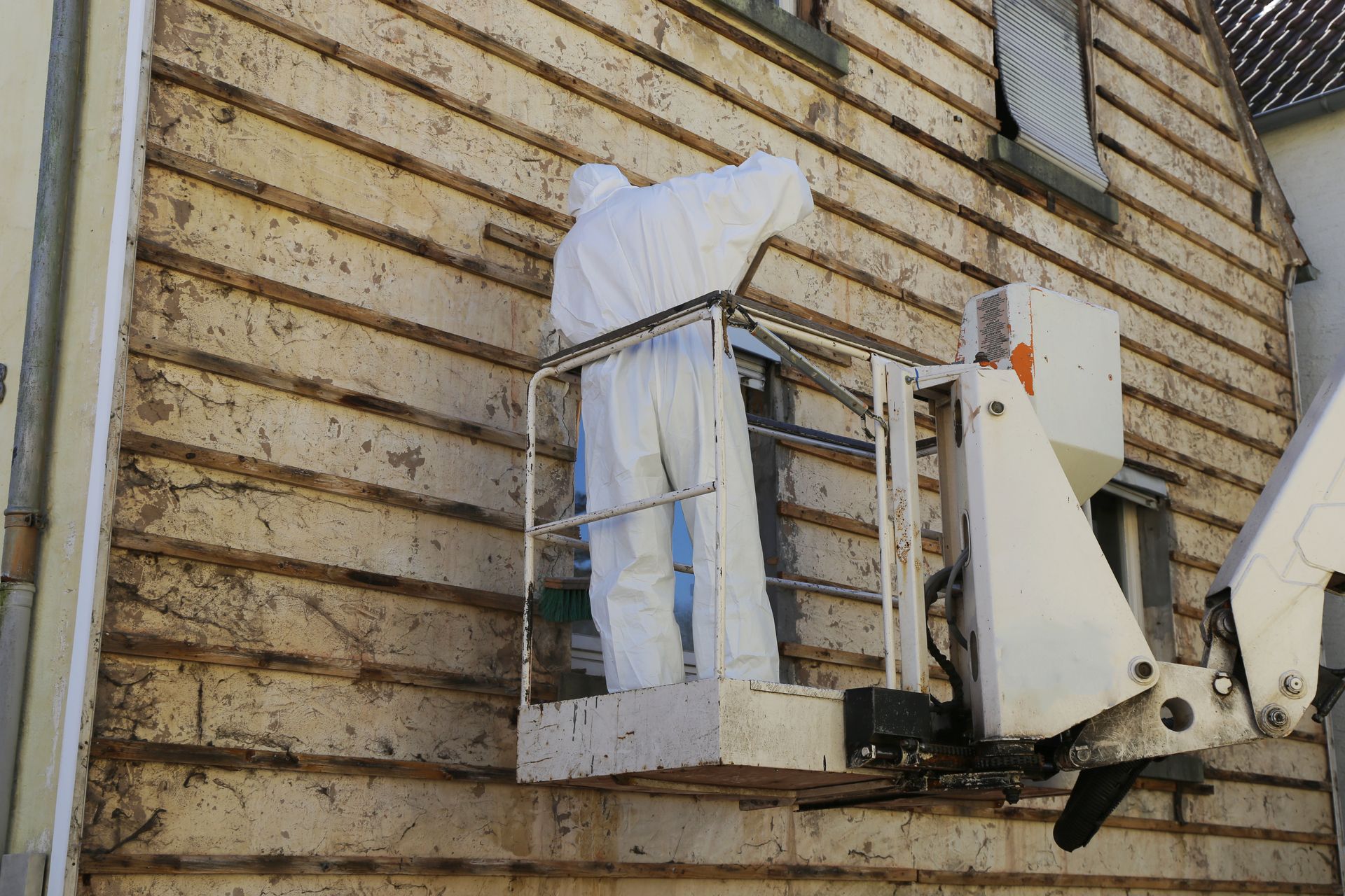 Worker in a white lift basket cleaning a brick wall with a high-pressure washer.