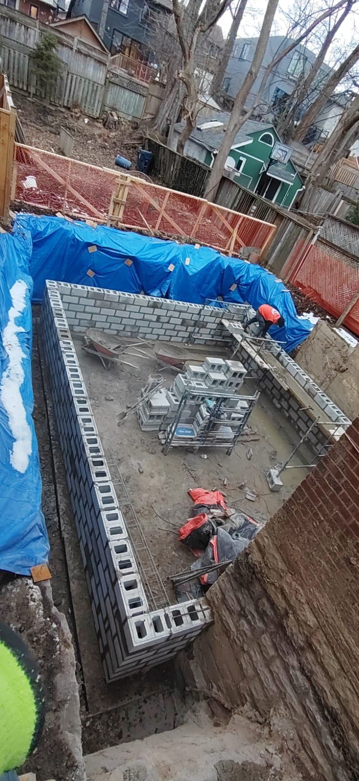 Construction site with a rectangular brick structure being built. A worker in an orange vest is visible. Blue tarp and safety fencing surround the site.