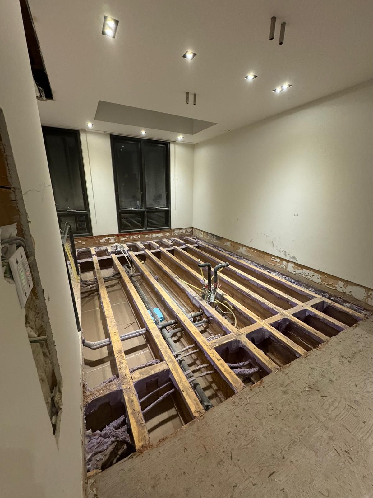 Room under construction with exposed floor joists, plumbing, and insulation. White walls and ceiling.