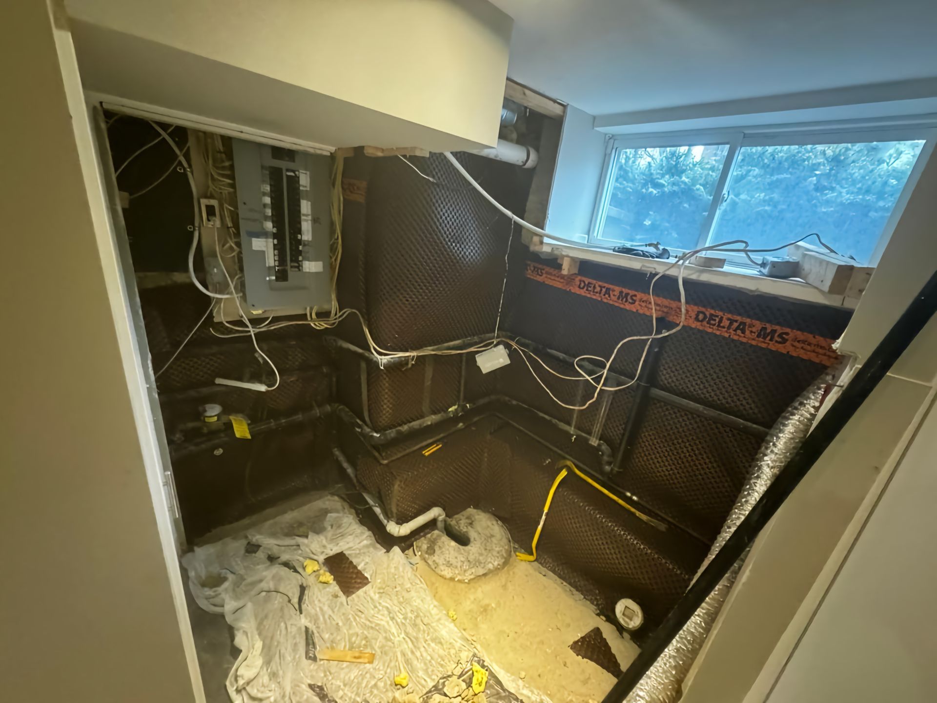 Basement renovation in progress; exposed electrical panel, walls with dark membrane, white wires, small window.