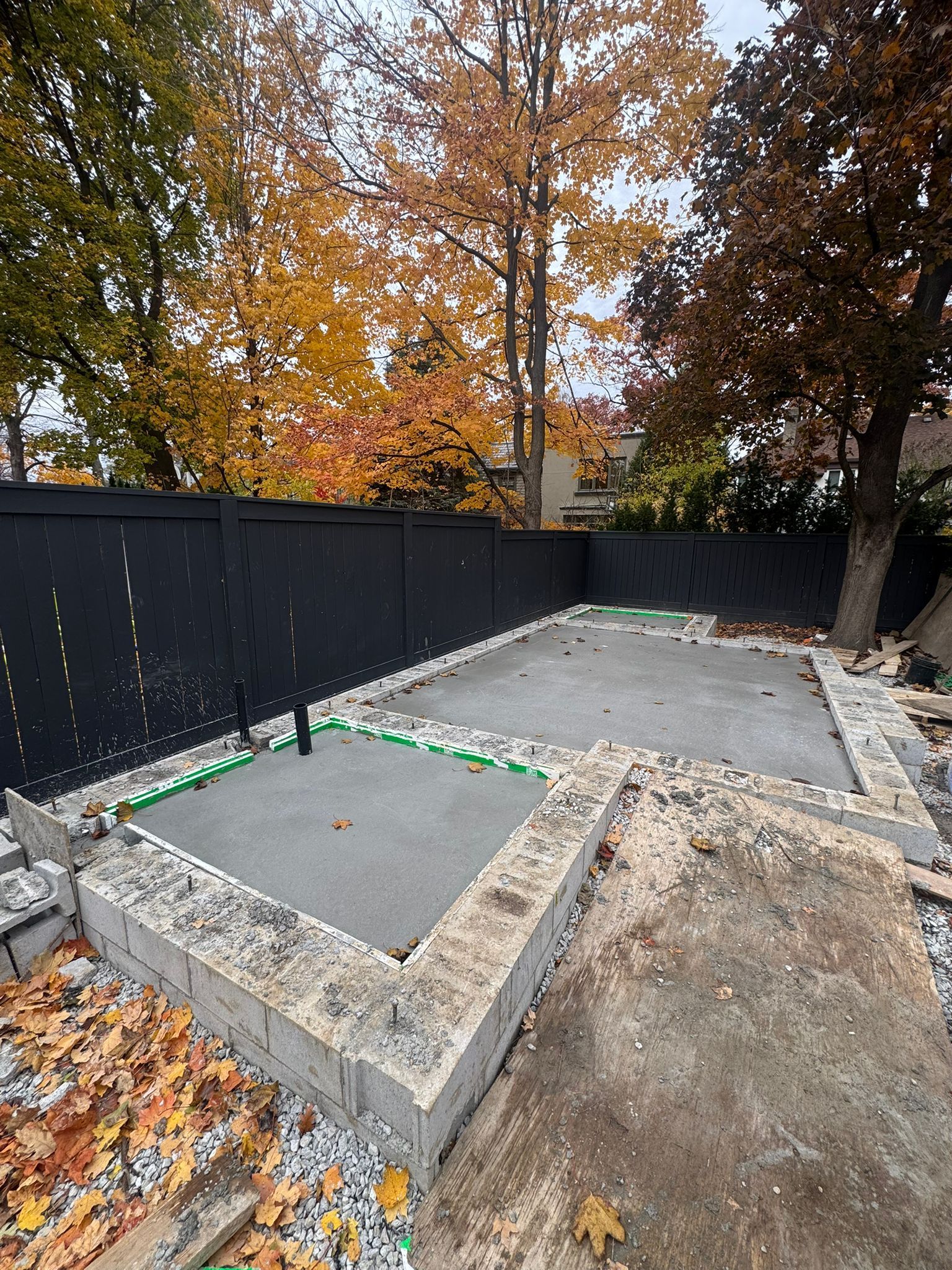 Concrete patio under construction with a black fence, surrounded by fallen autumn leaves.