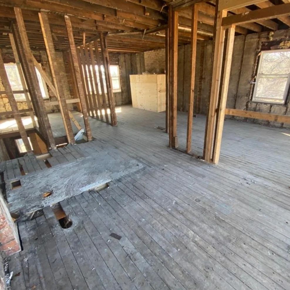 Interior view of a gutted, unfinished room with exposed wooden beams, studs, and worn wooden floors.