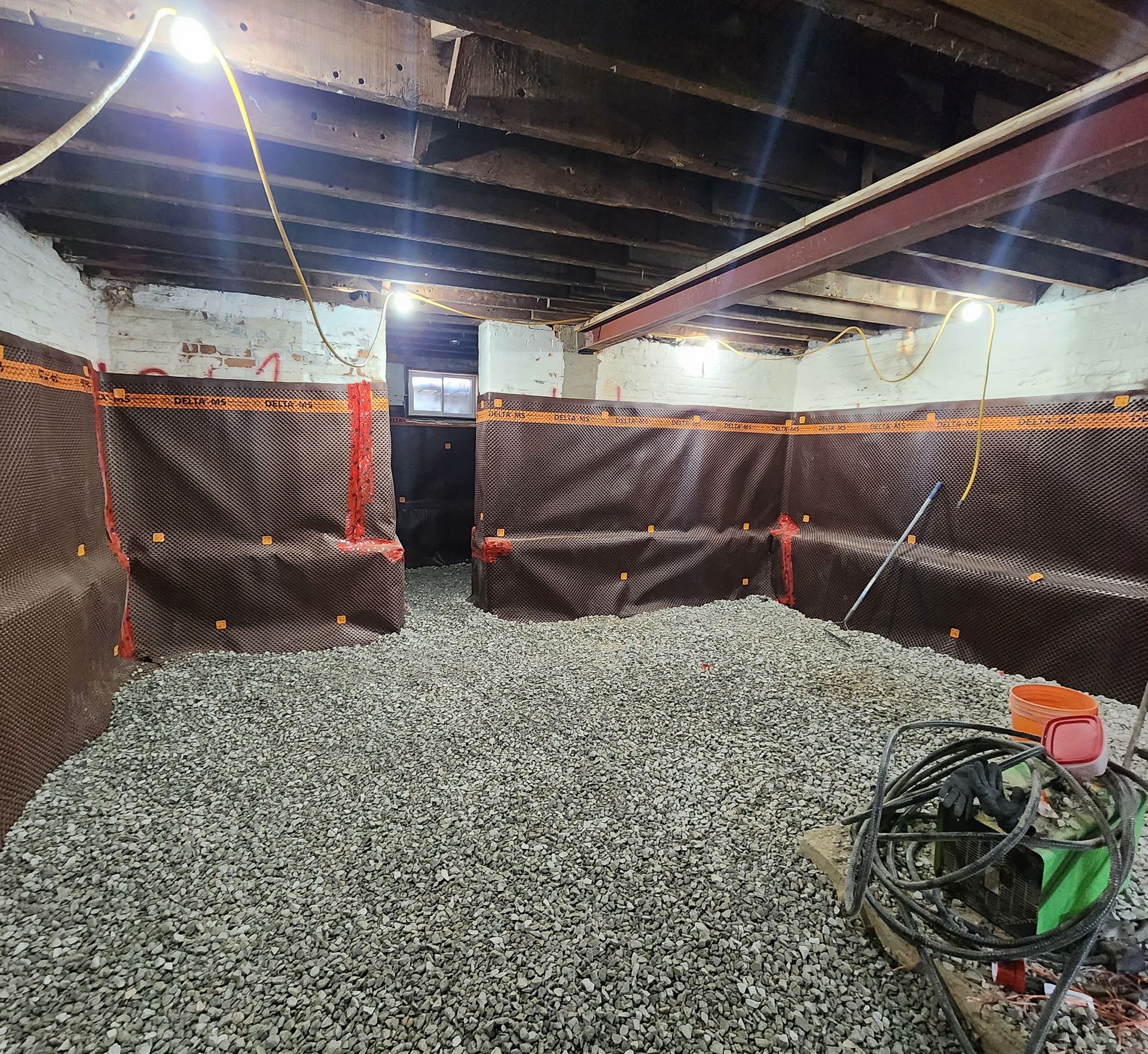 Basement renovation with gravel floor, dark walls with brown protective layer, exposed wooden beams, and lights.