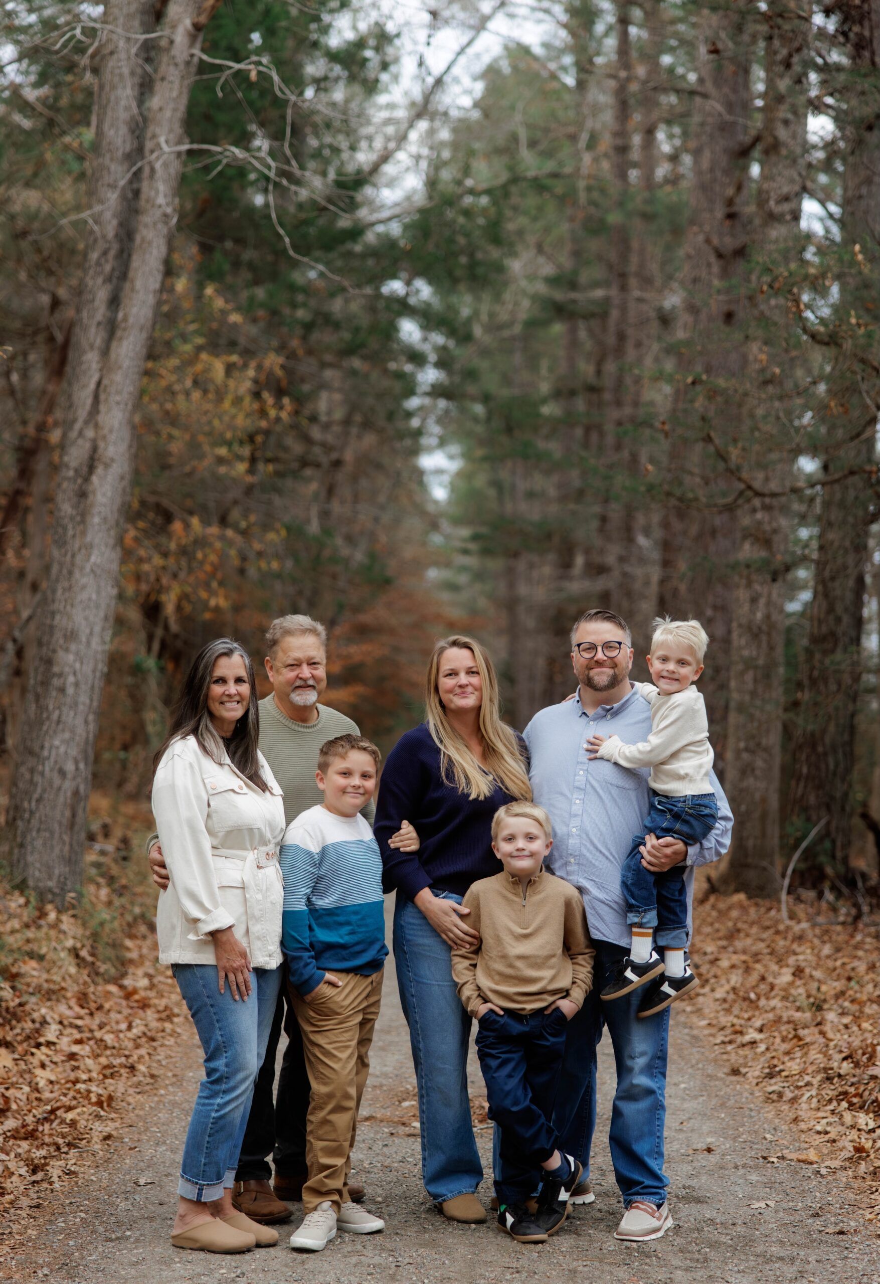 A multi-generational family poses together on a gravel path lined with pine trees during autumn.