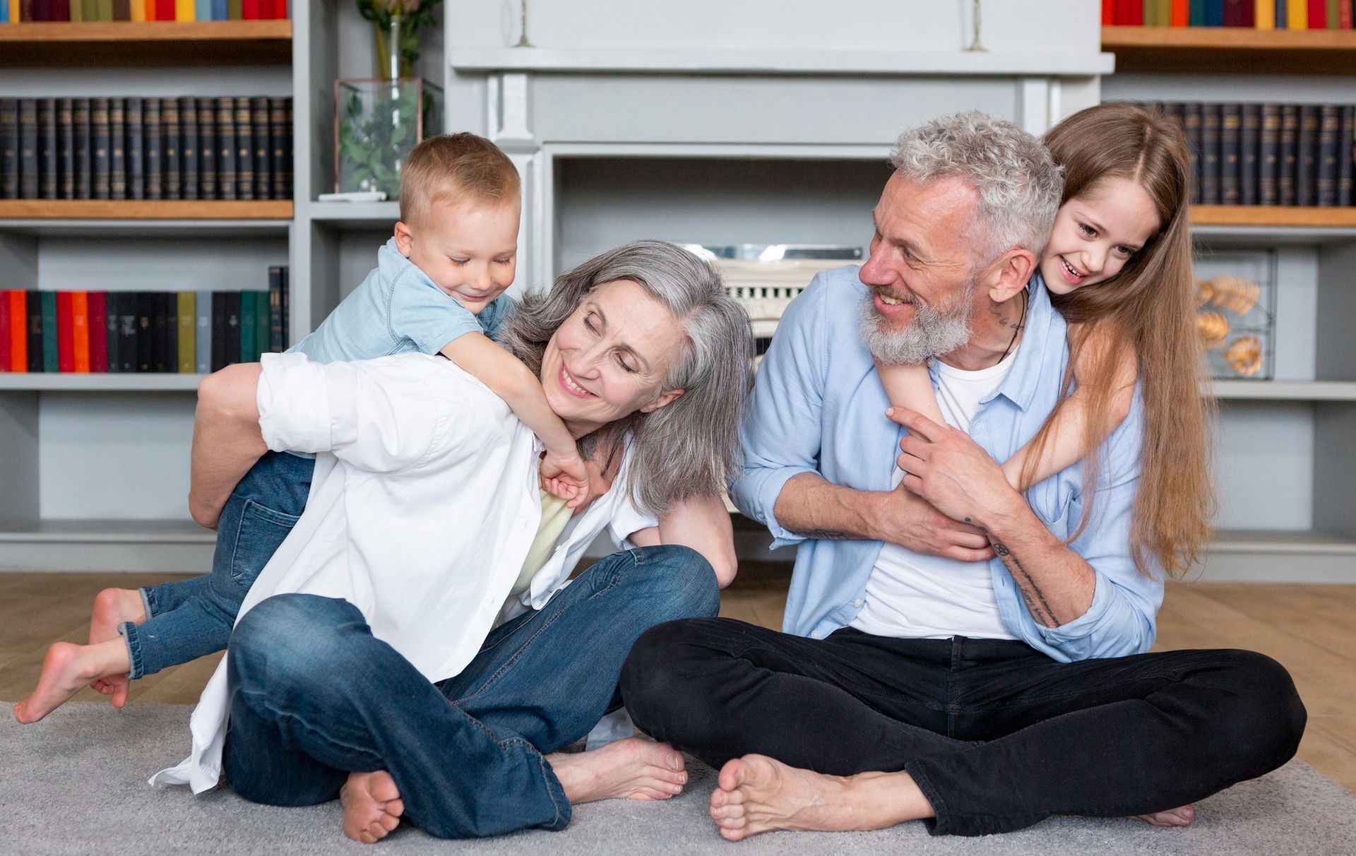 An older couple sits on a rug in front of bookshelves while children play on their backs, all smiling.