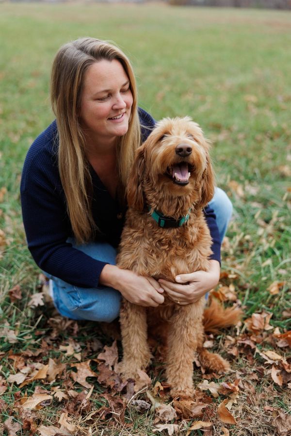 A person in a blue sweater and jeans kneels in a grassy, leaf-covered field, smiling while hugging a golden doodle dog.