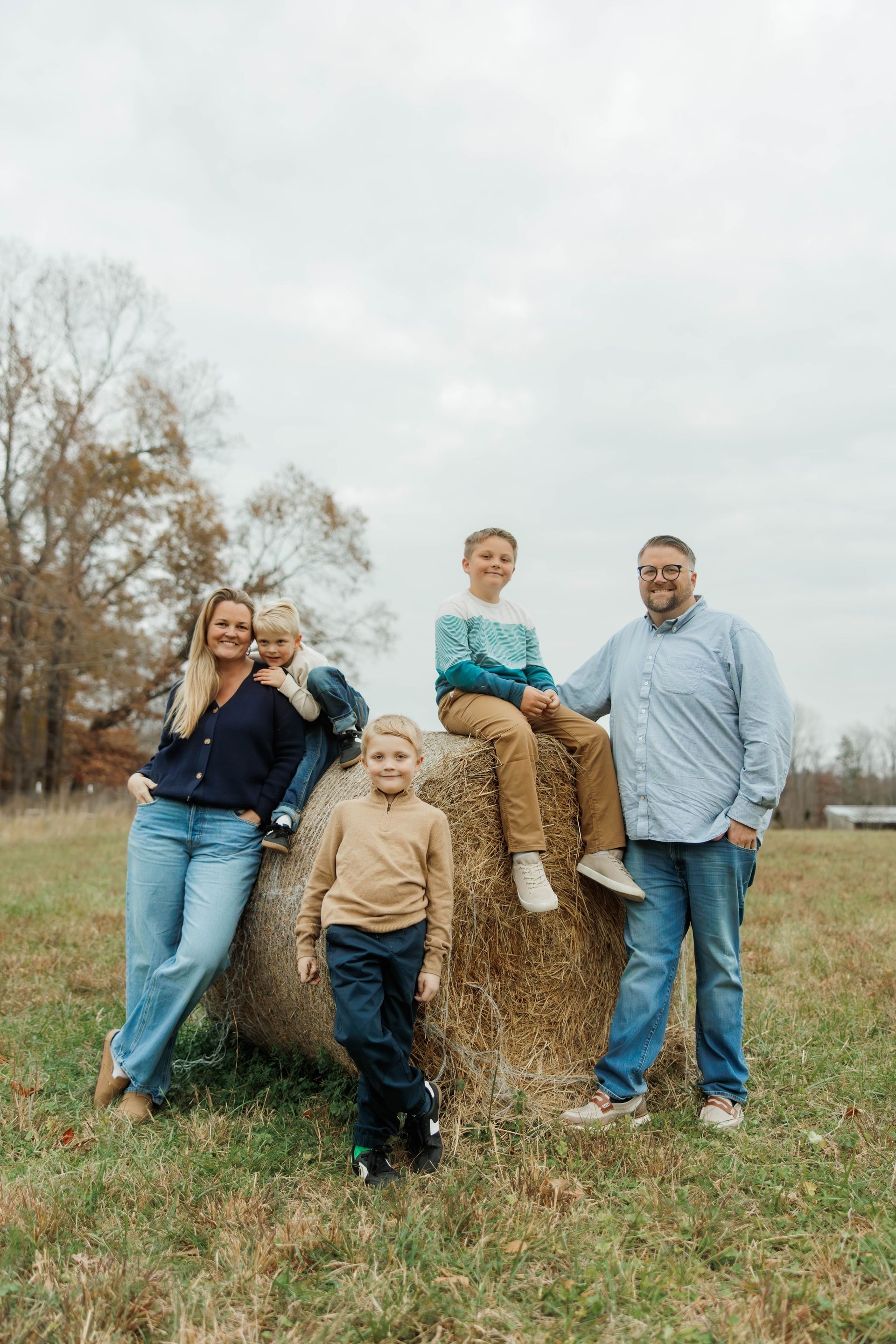 A family of five posing together in an autumn field by a large hay bale under a cloudy sky.