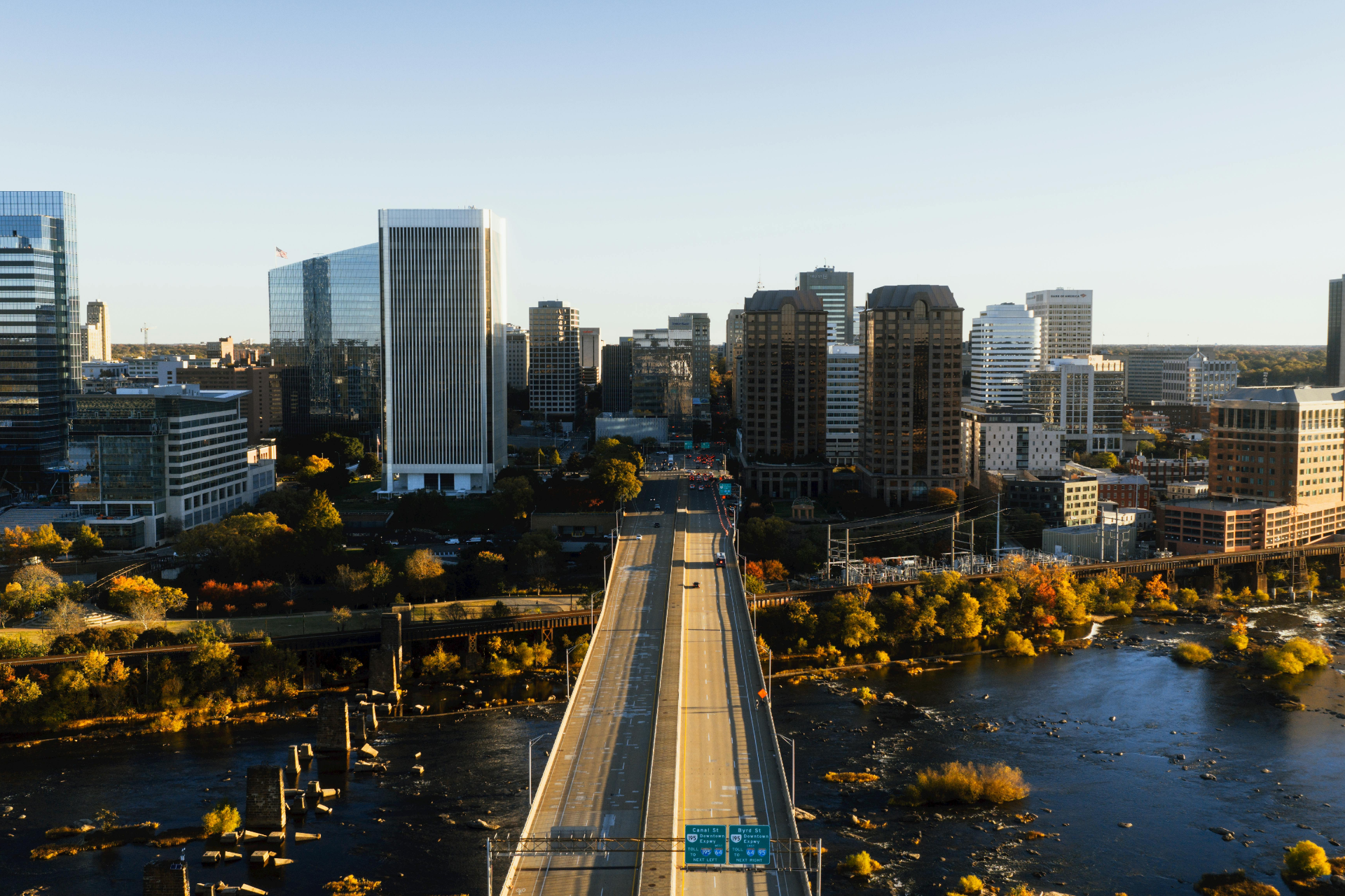 A bridge spans a river leading toward a city skyline under a clear, bright blue sky at sunset.
