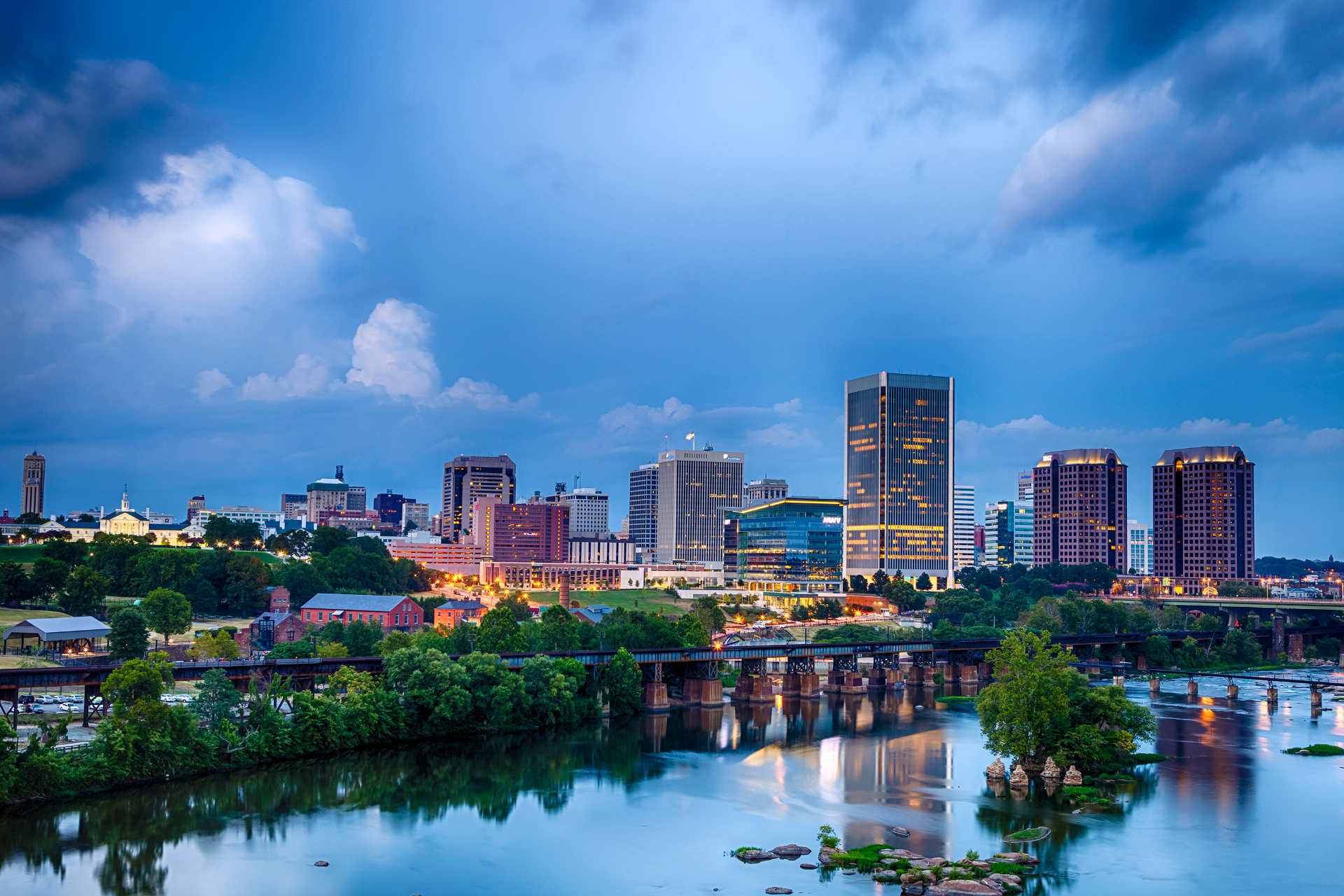 A scenic view of the Richmond, Virginia skyline at dusk, featuring city buildings reflected in the James River.