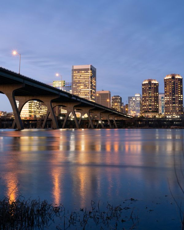 A city skyline at twilight reflected in a calm river, featuring a prominent arched bridge crossing the water.