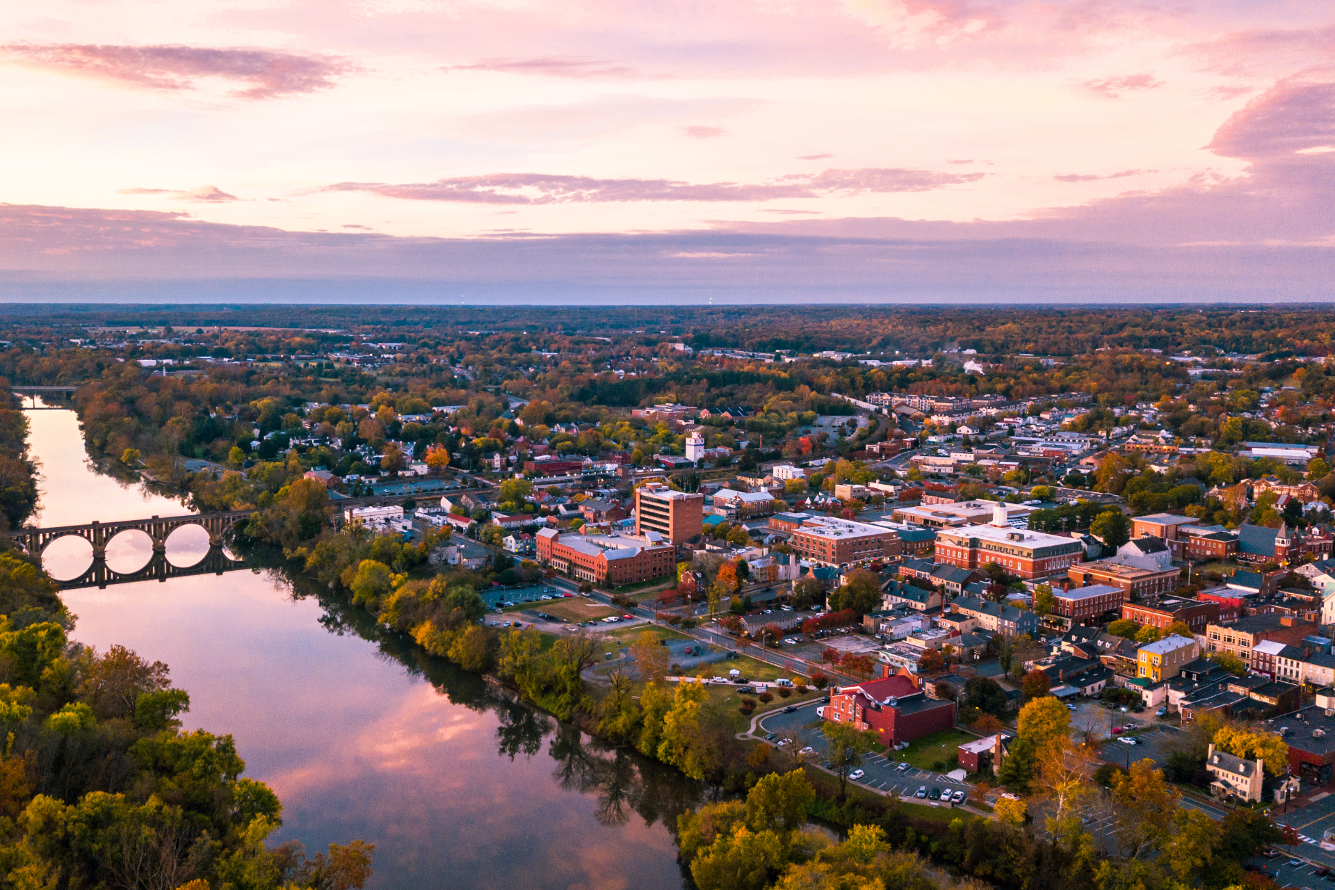 Aerial view of a small town at sunset, featuring a river with an arched stone bridge reflecting a pink and orange sky.