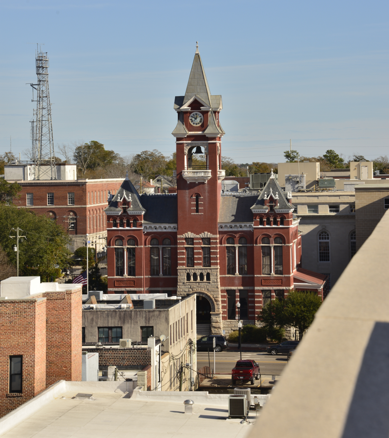 A historic red brick courthouse with a clock tower stands in a city square, with other buildings and a cell tower nearby.