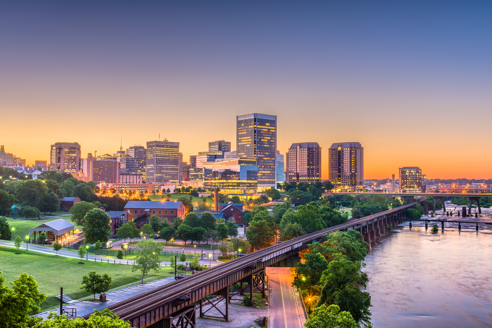 Sunset view of the Richmond, Virginia skyline over the James River and a historic railroad bridge.