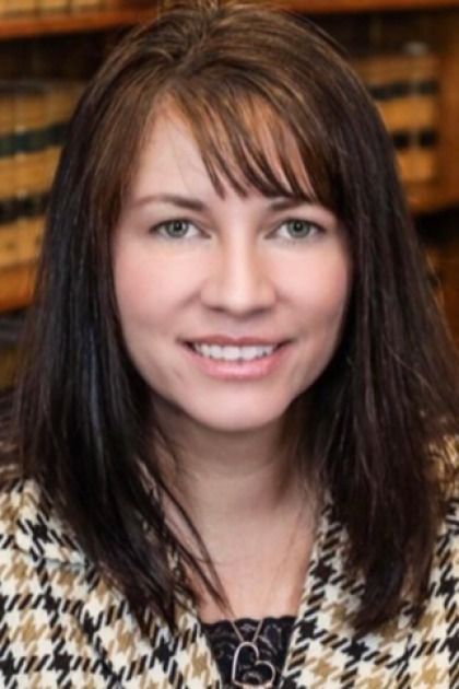 A person with long dark hair and bangs wearing a black and white houndstooth jacket in front of a bookshelf.