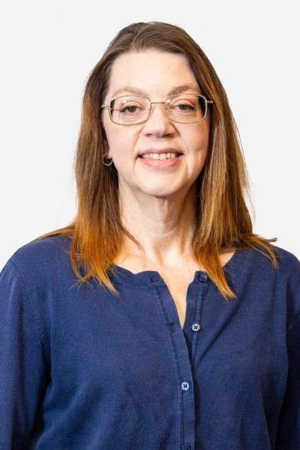 A smiling woman with brown hair and glasses wearing a blue collared shirt against a plain white background.