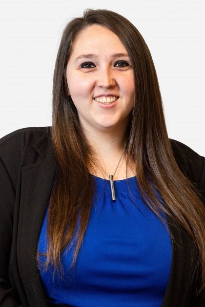 A person with long brown hair, wearing a royal blue top and a black blazer, smiling against a plain white background.