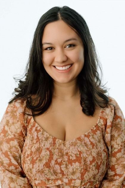 A person with dark hair smiling at the camera, wearing an orange and cream floral patterned top against a white background.