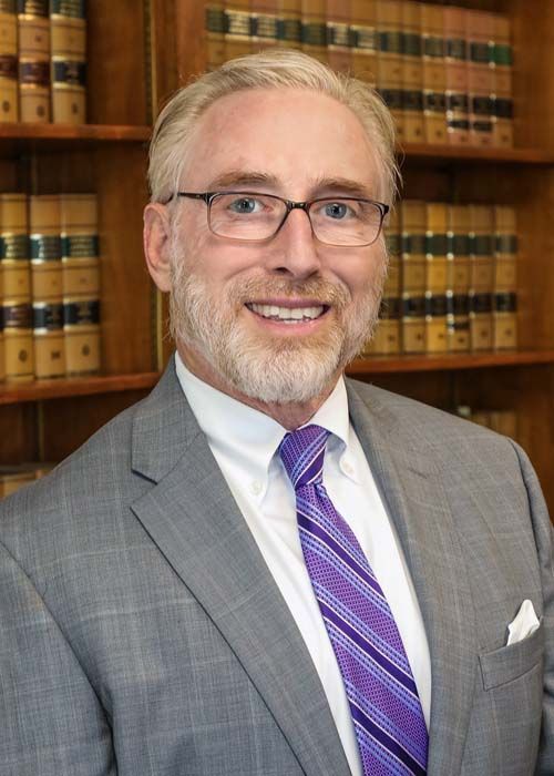 A professional portrait of a person in a gray suit and purple striped tie, smiling in front of a wooden bookshelf.