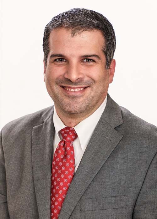 Headshot of a smiling person wearing a grey suit, white shirt, and patterned red tie against a white background.