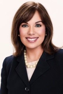A smiling professional with shoulder-length brown hair, wearing a dark blazer, light top, and pearl necklace.