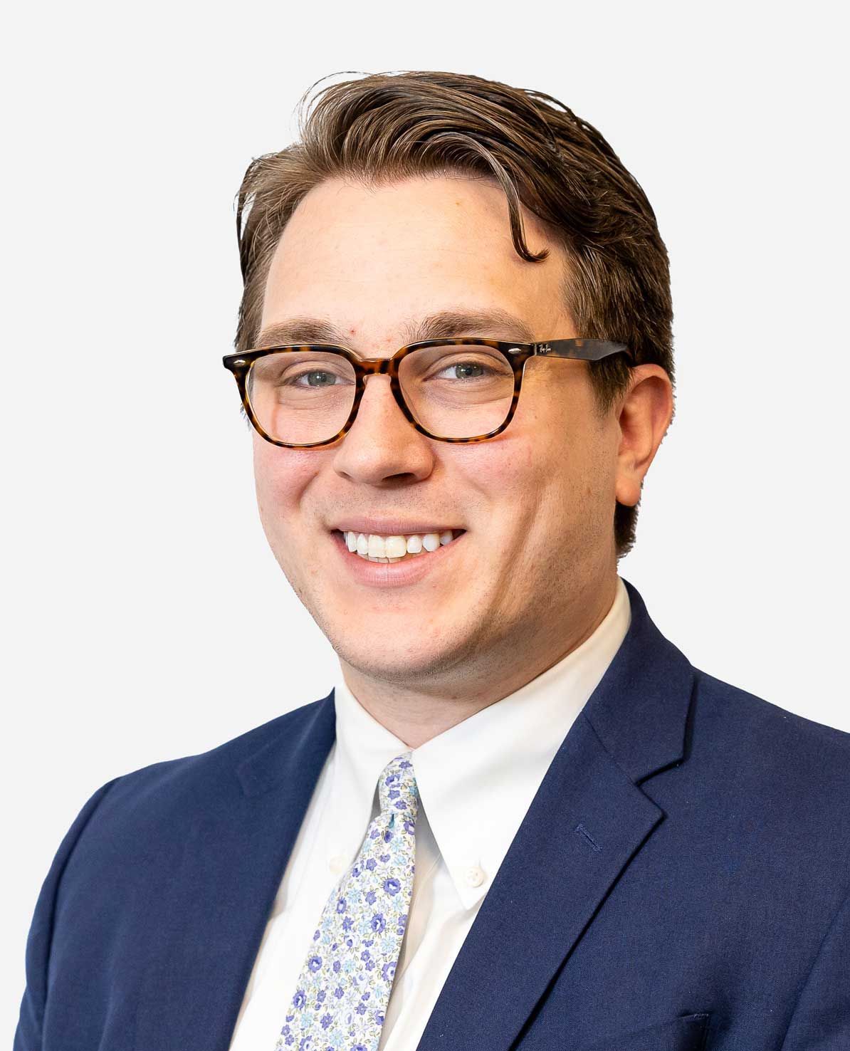 A person smiling, wearing glasses, a blue blazer, white shirt, and a light-patterned tie against a white background.