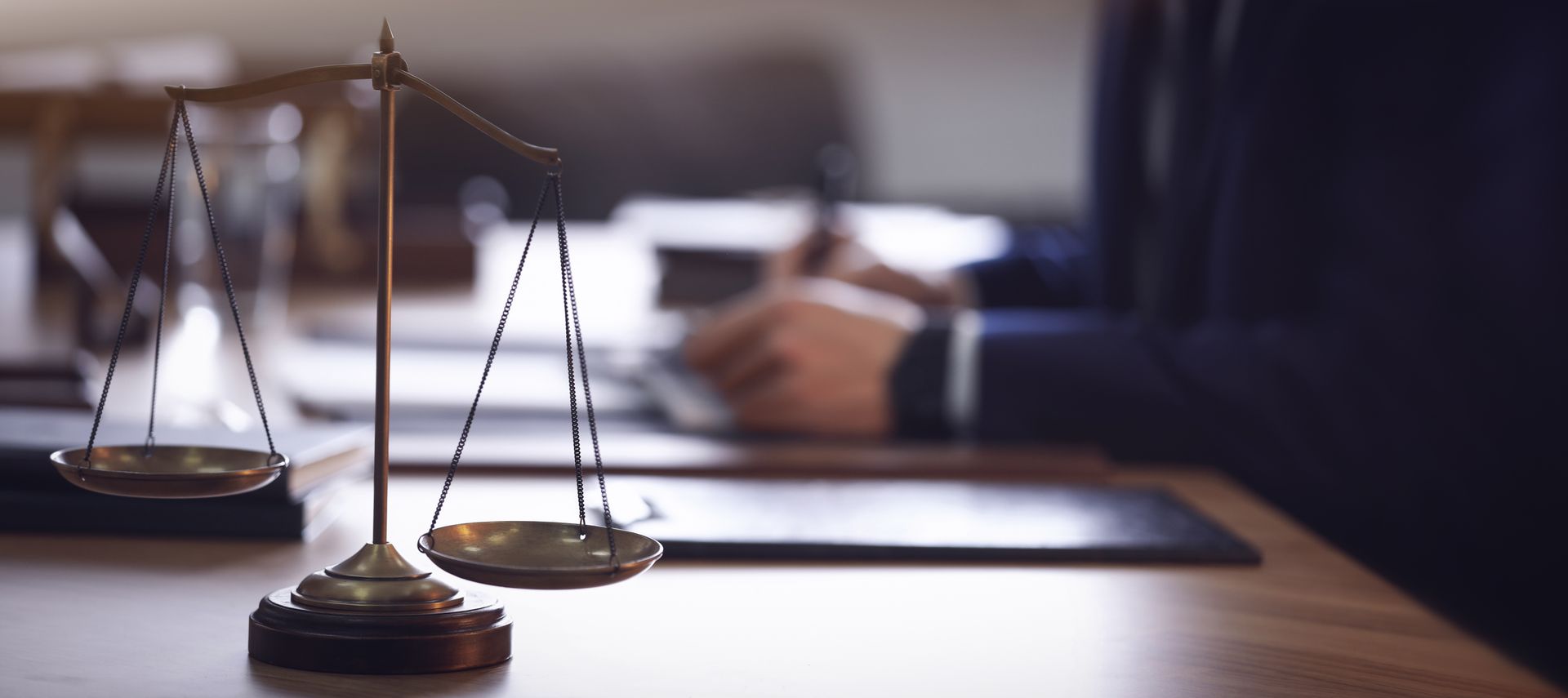 A brass scale of justice sits on a desk in the foreground, with a person in a suit working with documents in the background.
