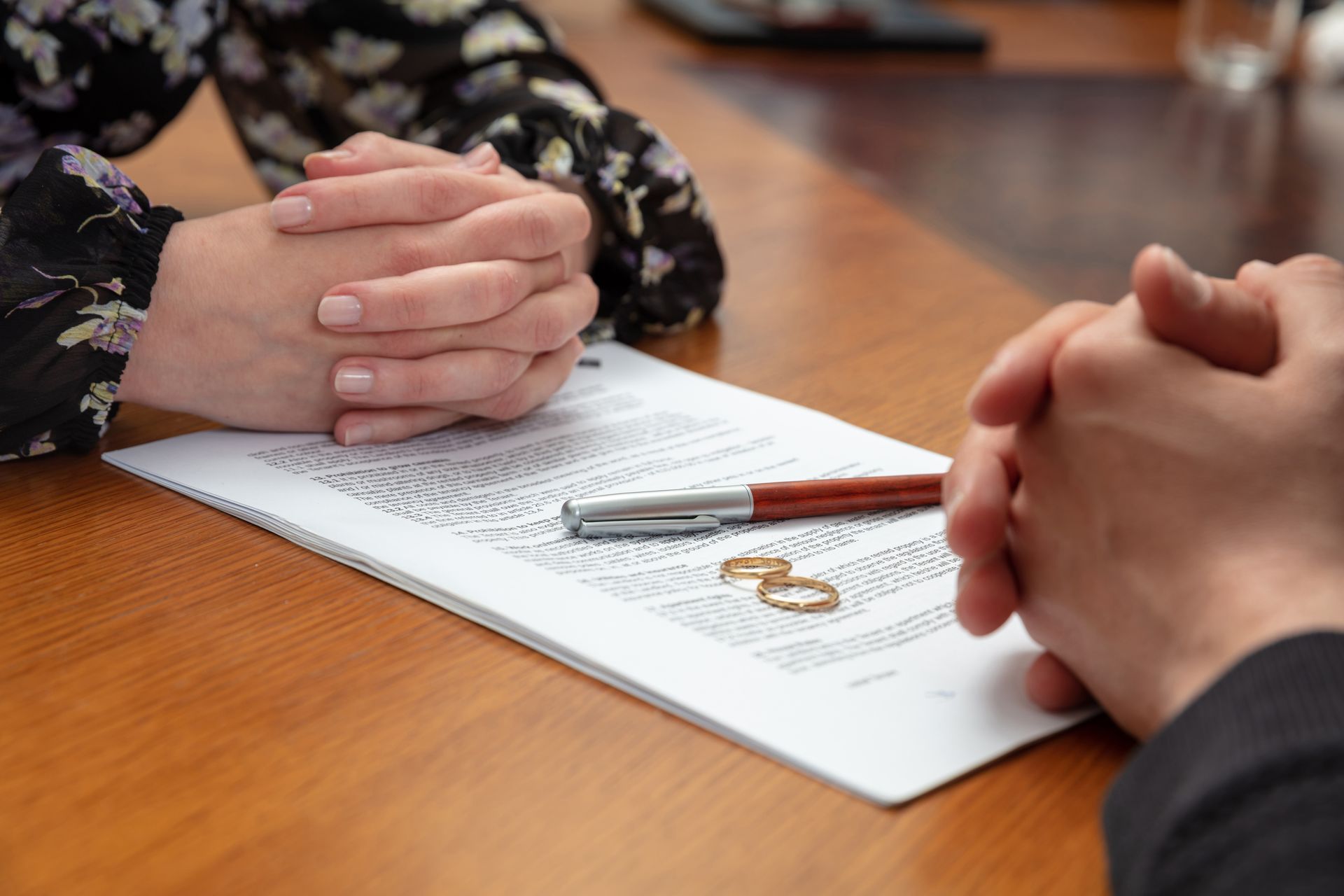 Two people sit at a wooden table with hands clasped over a legal document, a pen, and two wedding bands.