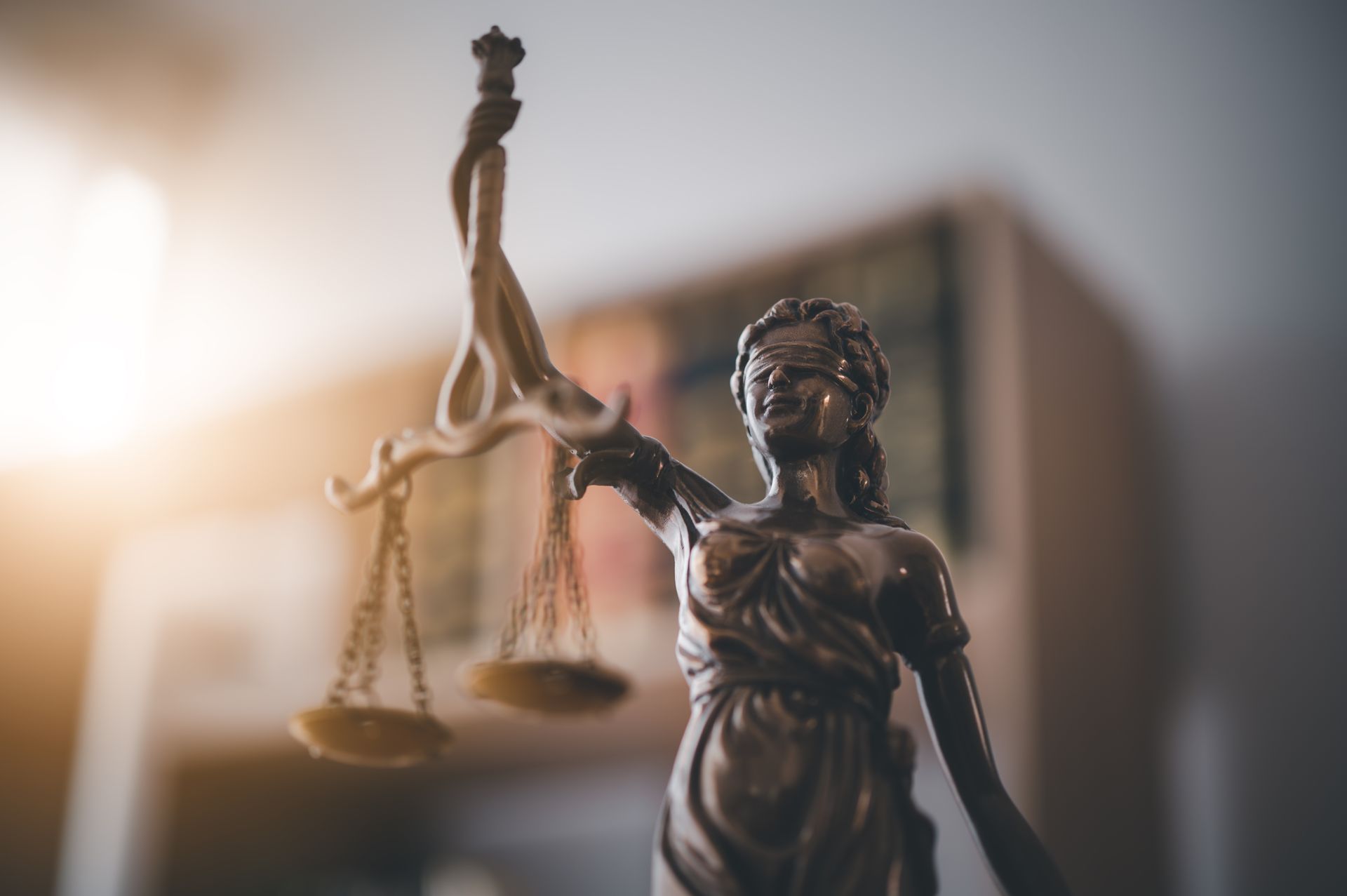 A bronze statue of Lady Justice holding scales, positioned in front of a blurred bookshelf in soft, warm lighting.
