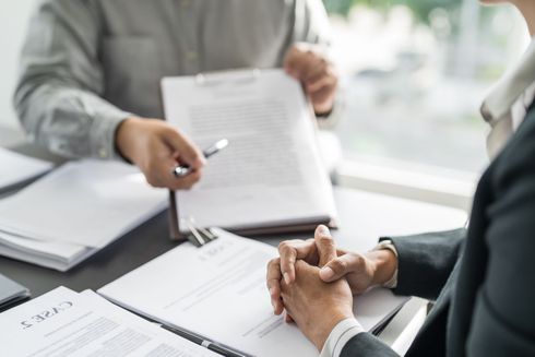 Two people in business attire sit at a table reviewing documents, with one person holding a pen over a contract.