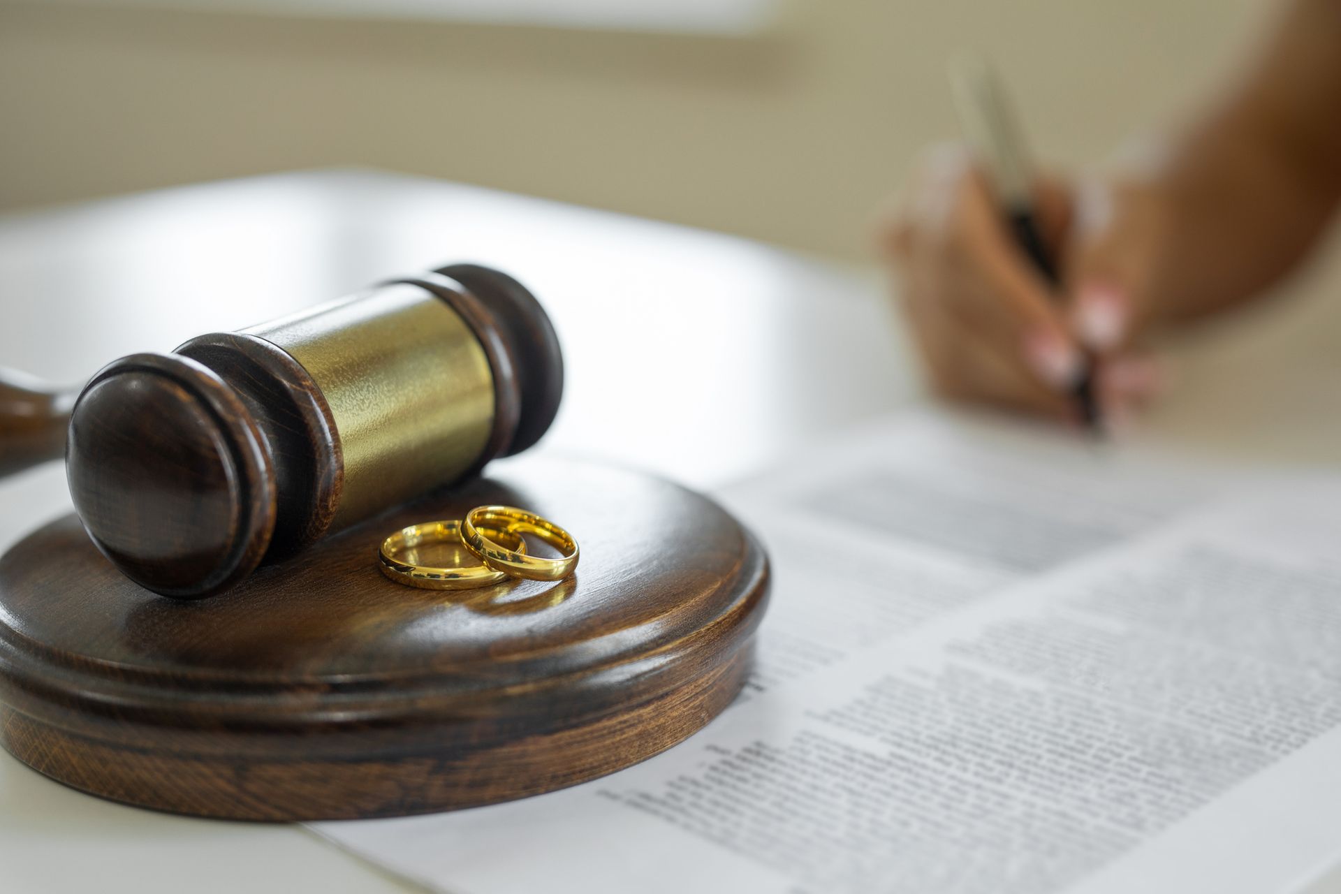 Wedding rings and a wooden gavel with a person signing a contract at a divorce law firm office.