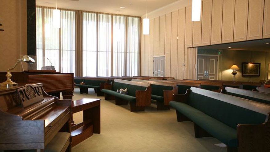 Empty chapel with wooden pews, large windows, and soft daylight.