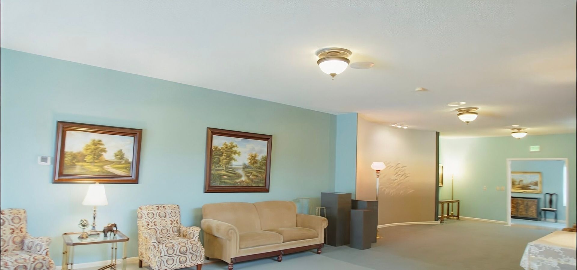 Bright lounge with floral chairs, beige sofa, framed art, and soft blue walls in a spacious hallway.