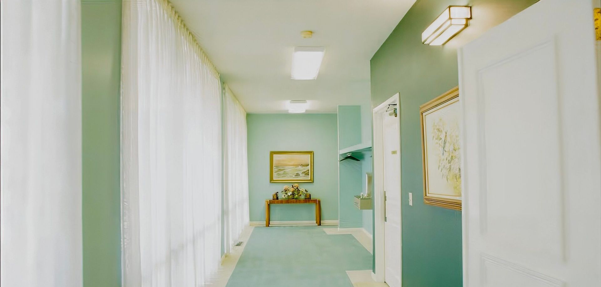 Narrow pastel hallway with white curtains, teal walls, and a small table with framed art at the far end
