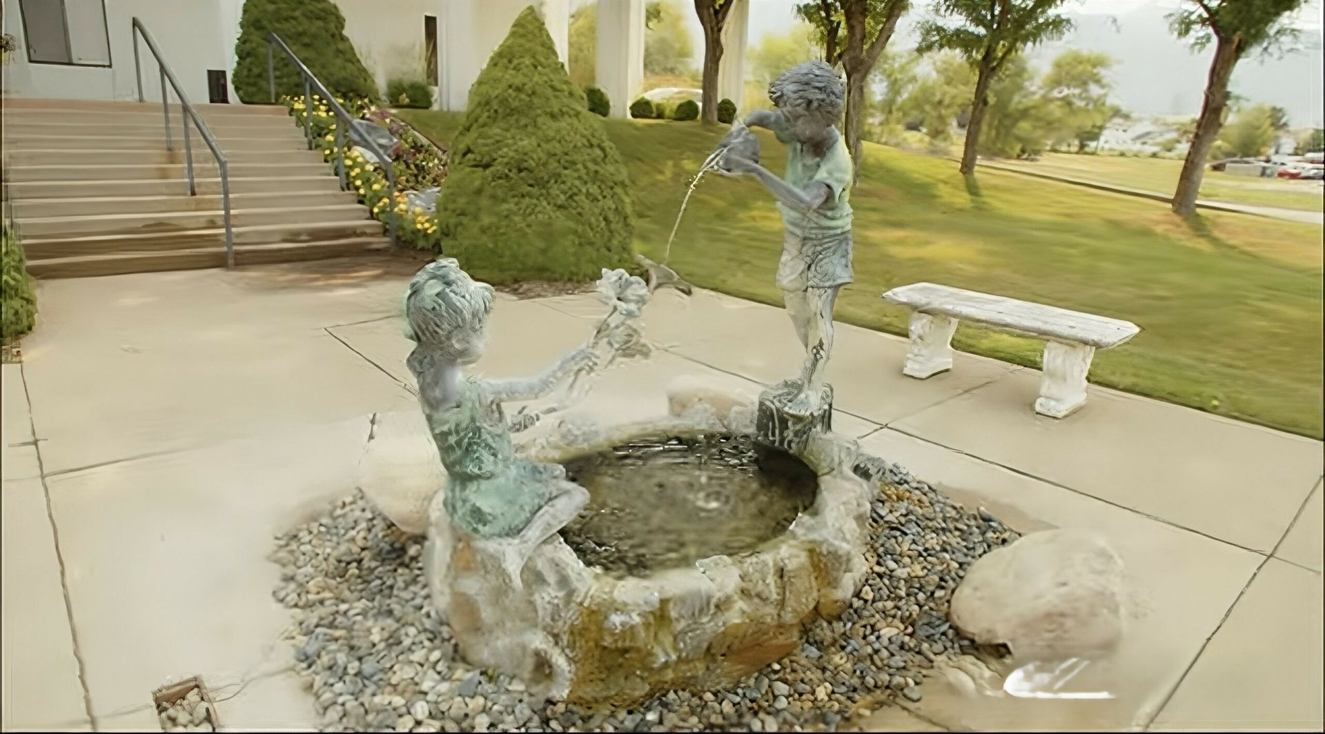 Bronze child statues around a small stone fountain in a park courtyard.