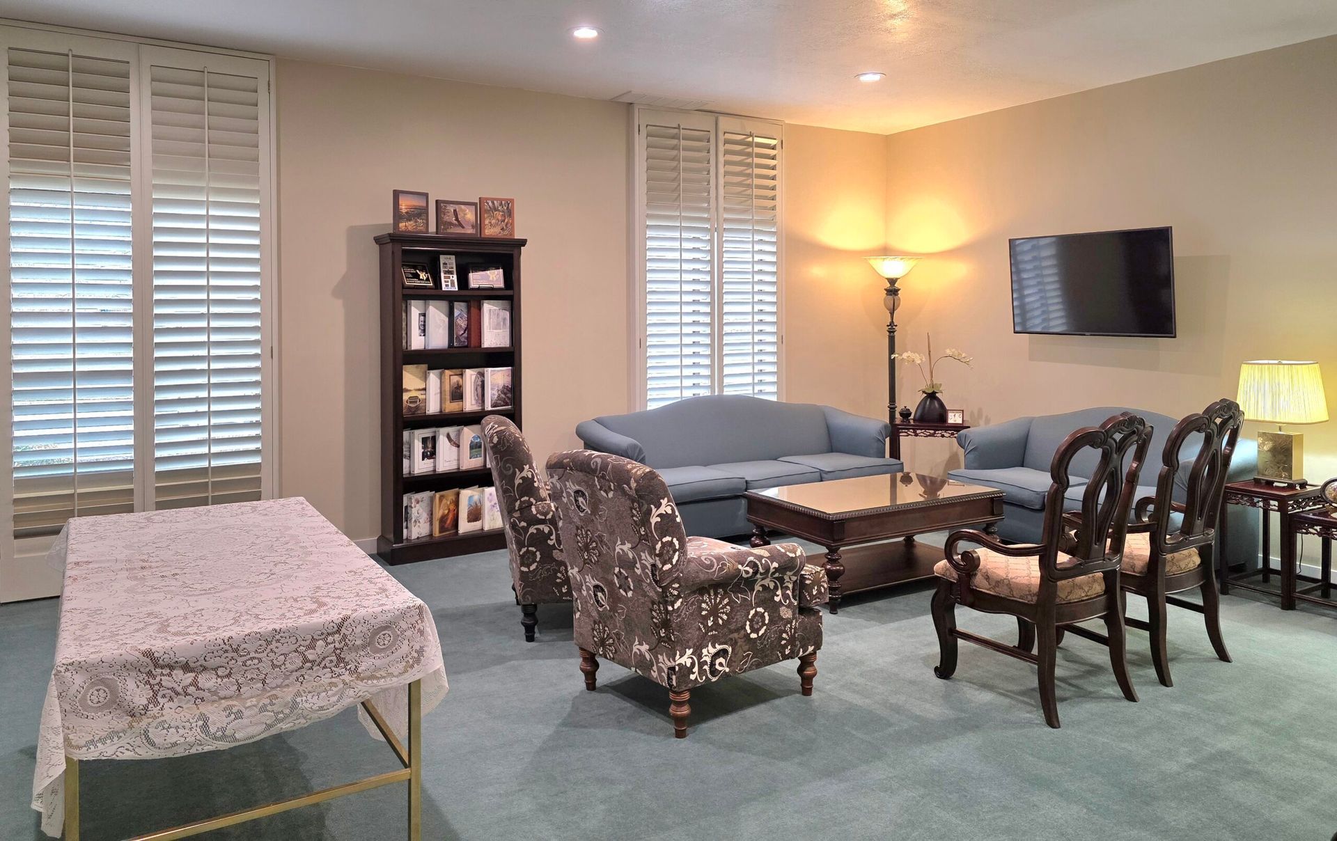 Spacious living room with green carpet, floral furniture, bookshelf, wall TV, lamps, and tall windows with blinds