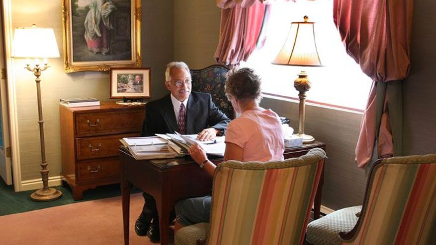 Two people seated in a cozy living room, having a conversation near a bright window and lamp.
