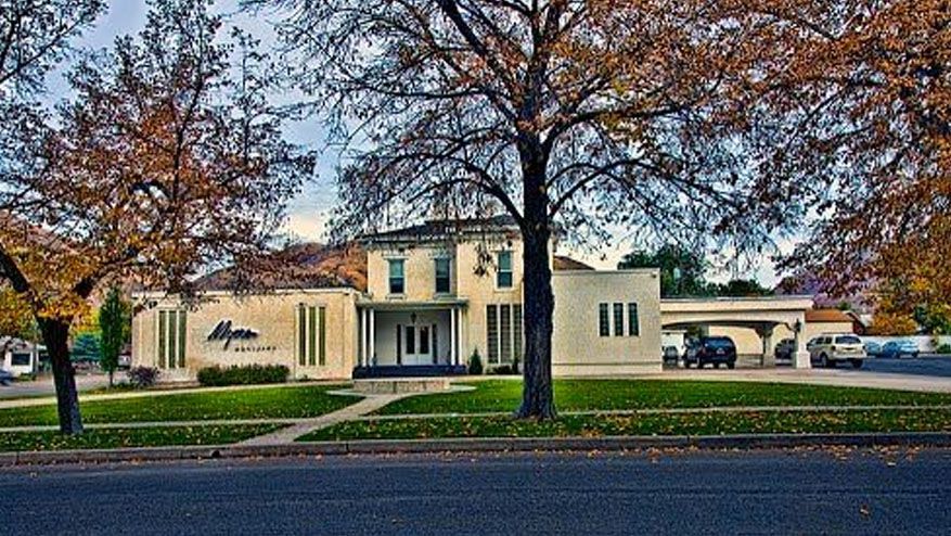 Autumn trees frame a cream-colored building with a central entrance and parked cars along the street.
