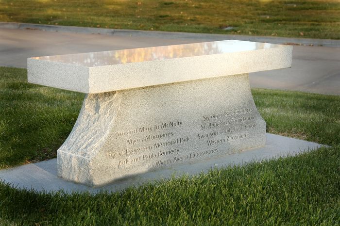 Stone memorial bench with engraved inscription on a grassy lawn near a road