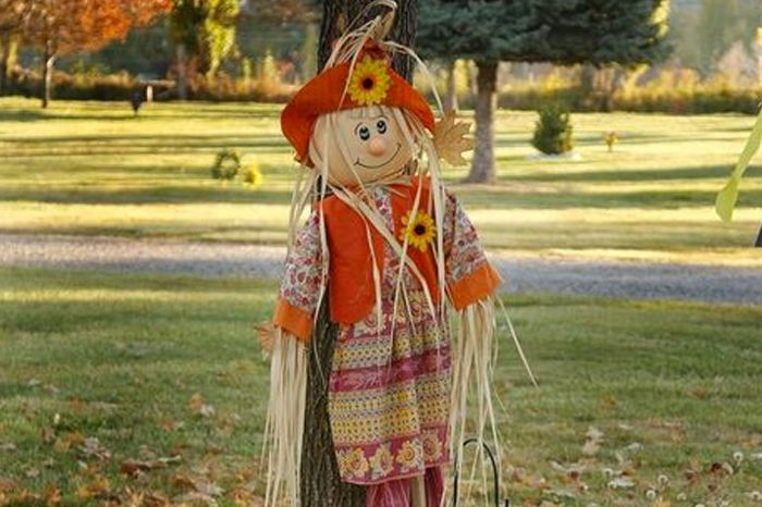 Scarecrow-like doll in orange-red clothes standing in a grassy park with autumn trees.