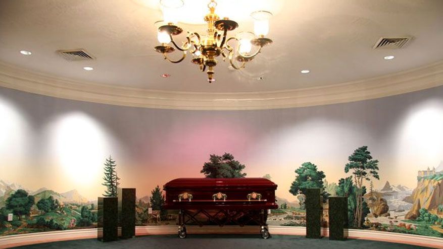 Indoor memorial hall with a red casket, floral arrangements, murals, and a chandelier