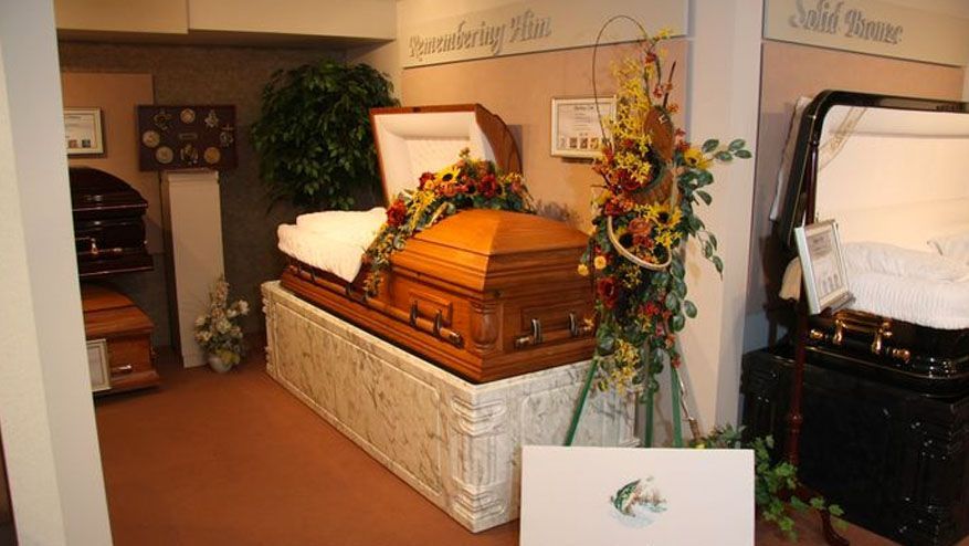 Casket in a funeral home viewing room, surrounded by flowers and a framed memorial display.
