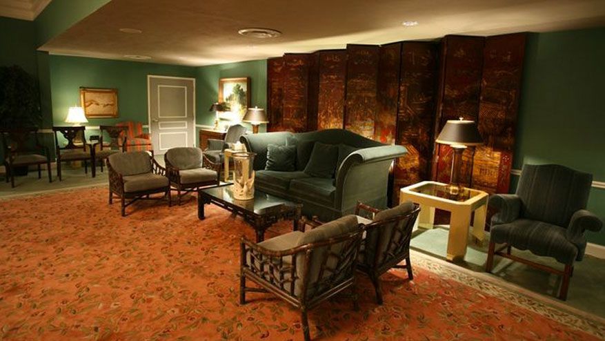 Cozy living room with green sofas, patterned chairs, lamps, and red carpet under warm lighting