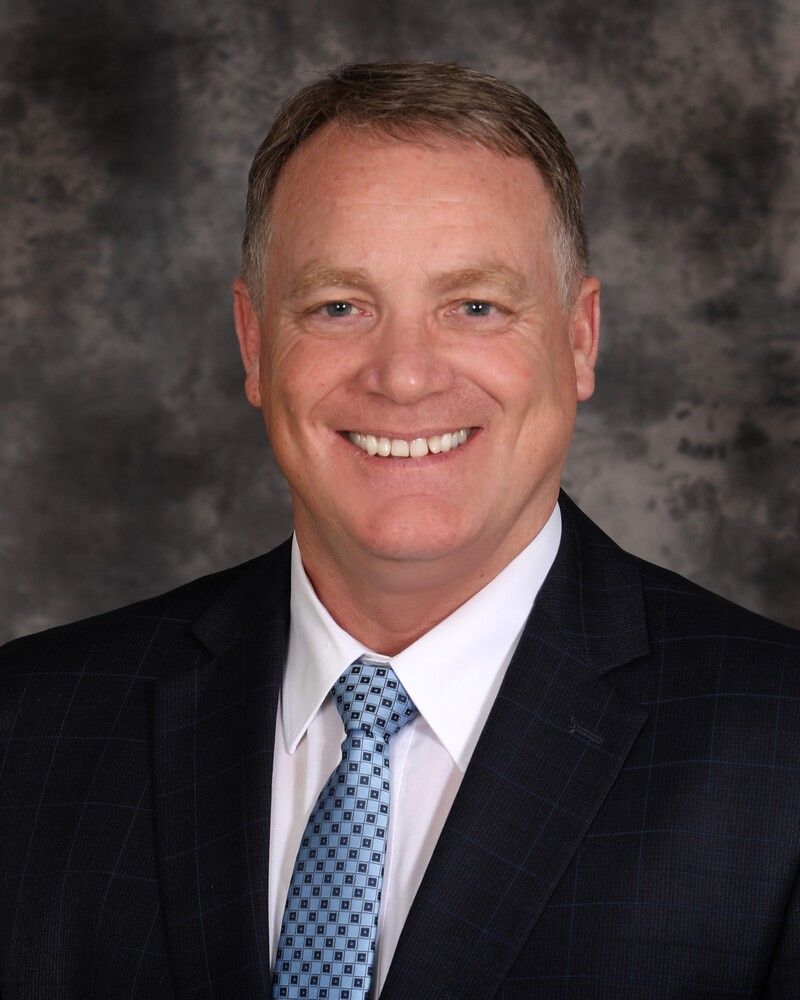 Smiling man in dark suit and blue tie against a gray studio backdrop