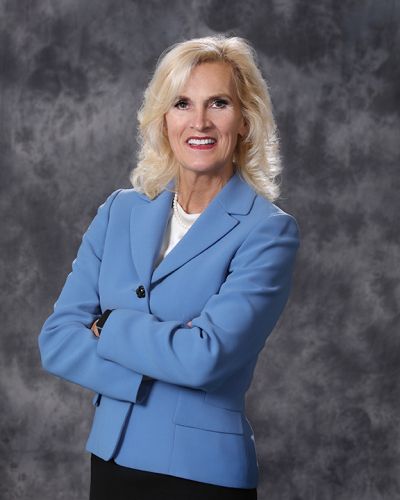 Blonde woman in a light blue blazer stands with arms crossed against a gray backdrop.