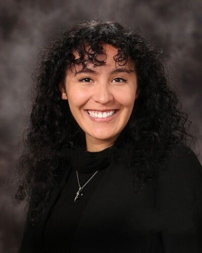Smiling person with curly dark hair wearing a black top and silver necklace against a gray studio backdrop